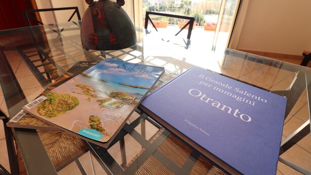 Una mesa de vidrio con dos libros titulados 'Grande Salento' y 'Otranto' y un cerdito de cerámica decorativo.