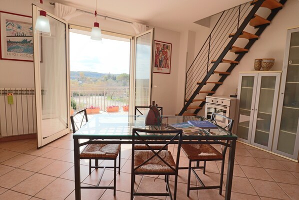 Un coin repas avec une table en verre, quatre chaises, une vue à travers la porte vitrée, une cuisine avec des placards et un escalier en bois.
