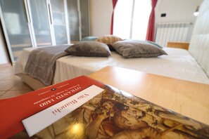 Un lit avec des oreillers, une couverture et un livre sur une table en bois, dans une pièce dotée d’une fenêtre et de rideaux.