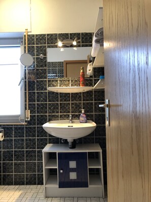 Une salle de bain avec un lavabo blanc, un miroir et un porte-serviettes. Il y a une fenêtre donnant sur l’extérieur.