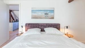 A bedroom with a large bed, a painting of a beach scene, and a view of a kitchen and dining area. - Vacation home Arlau Schleuse in the Hattstedtermarsch for up to 4 people (Hattstedtermarsch)