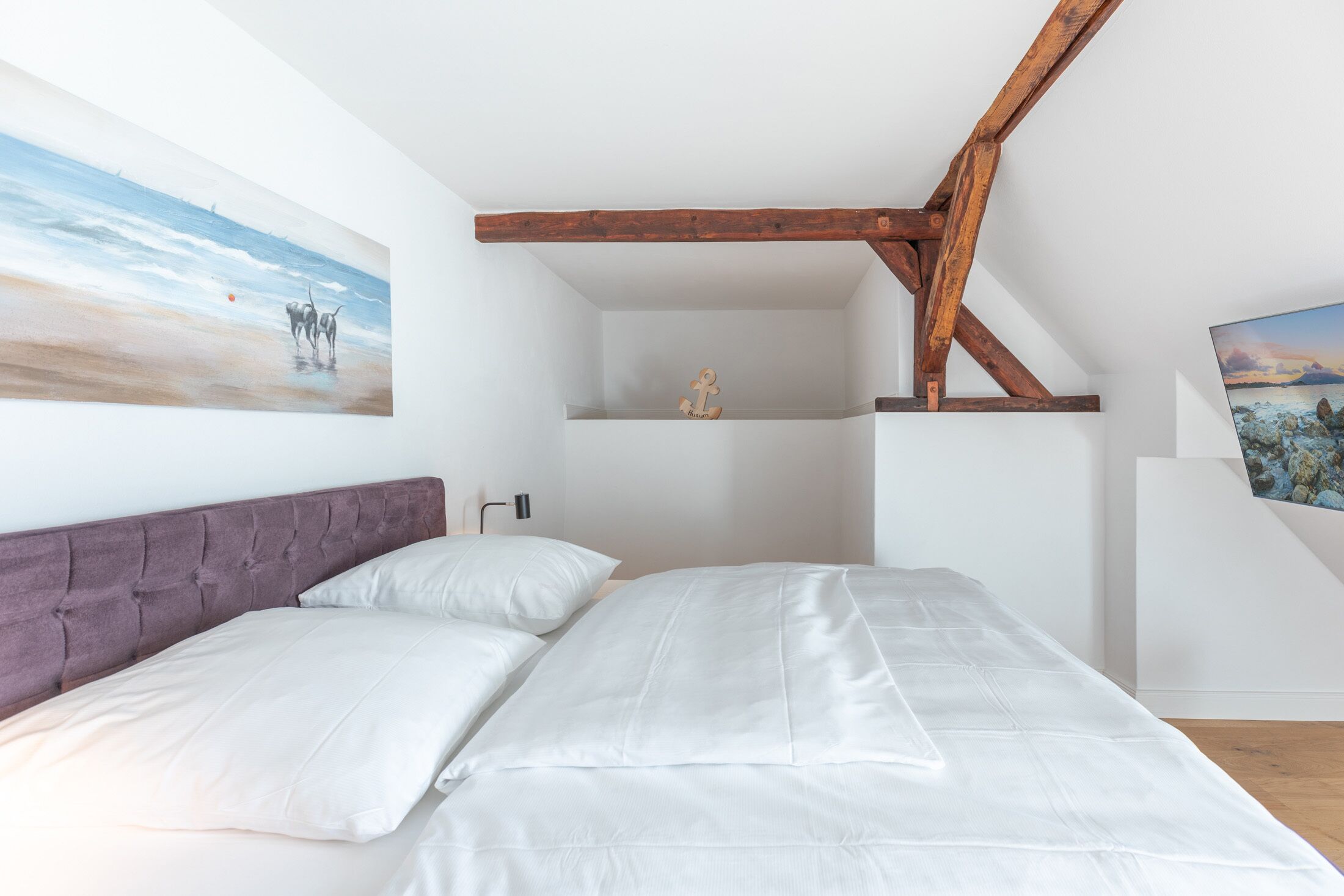 Une chambre à coucher avec un lit, du linge de lit blanc, une tête de lit violette, des poutres en bois et deux tableaux accrochés aux murs.