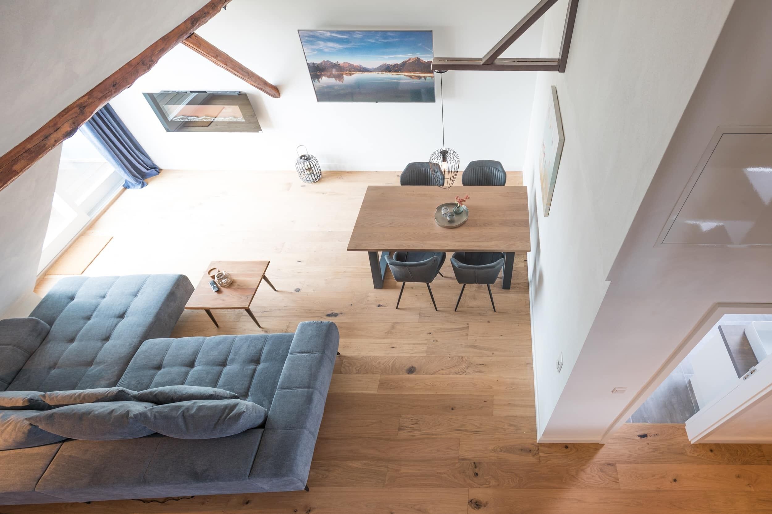 Een loft-achtige kamer met een houten vloer, een grijze bank, een eettafel met stoelen en een groot raam met uitzicht op een meer.