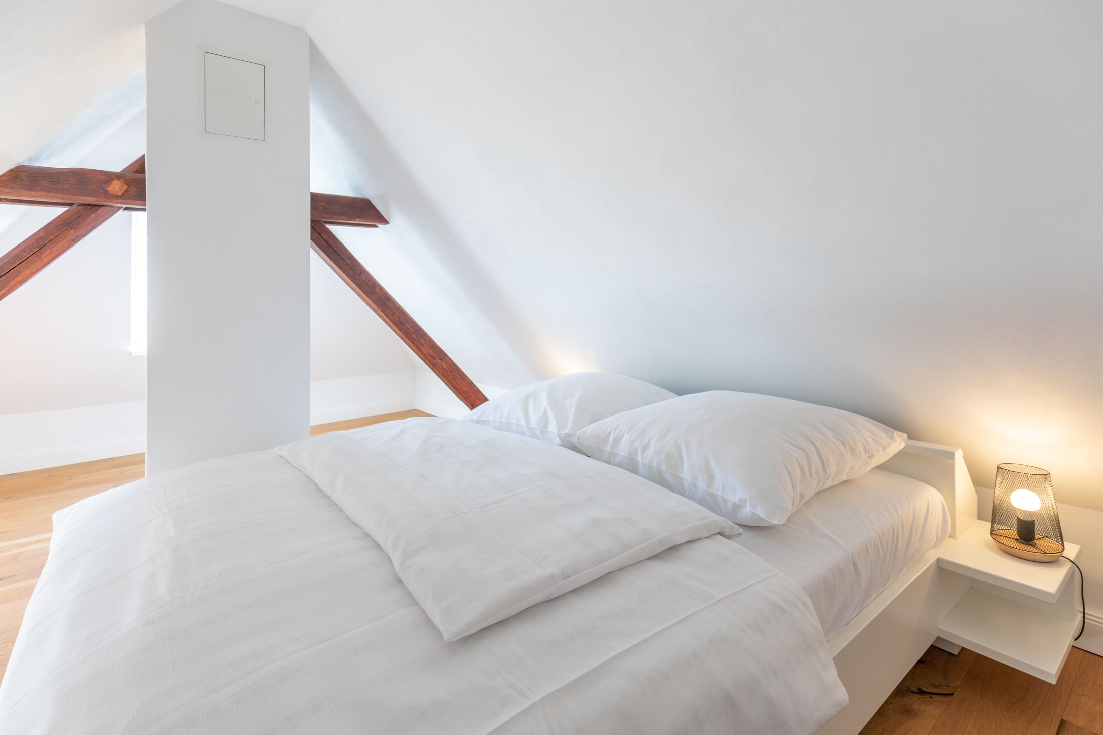 A bedroom with a sloped ceiling, a bed with white bedding, a bedside table with a lamp, and wooden beams.