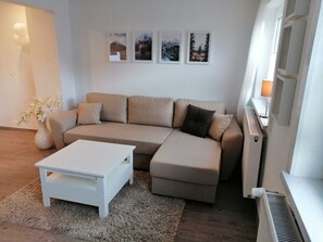 A modern living room with a beige sectional sofa, a white coffee table, and a small side table. There are framed pictures on the wall and a radiator on the right. - Knebel vacation apartment (Clausthal-Zellerfeld)