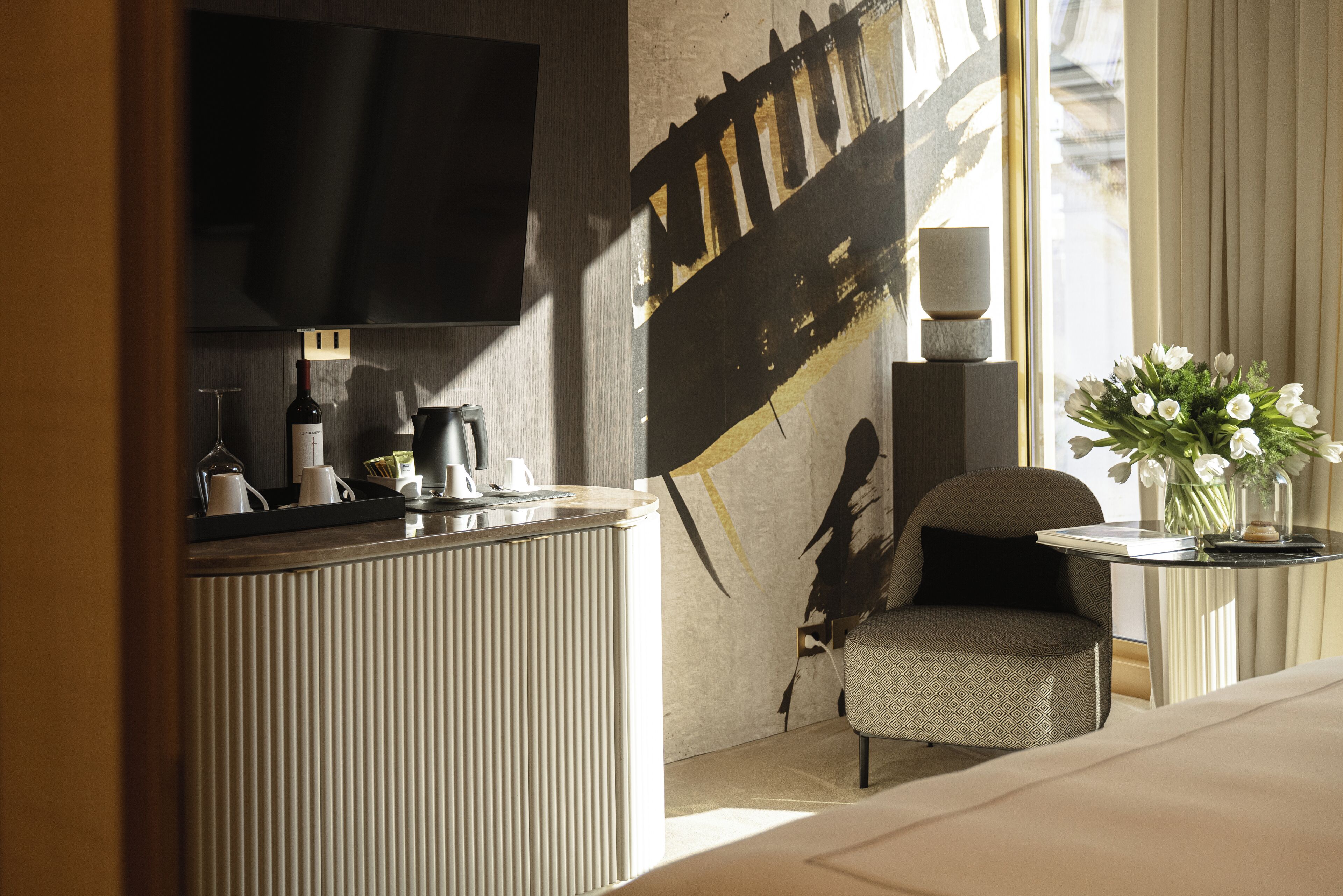 Een hotelkamer met een bed, een bureau met een flatscreen televisie, een stoel, een vaas met bloemen en een muur met een groot abstract schilderij.
