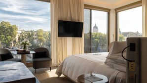 A hotel room with a large bed, a TV mounted on the wall, a view of a cityscape, and a small table with a book and a vase of flowers. - The First Musica - Preferred Hotels & Resorts (Rome)