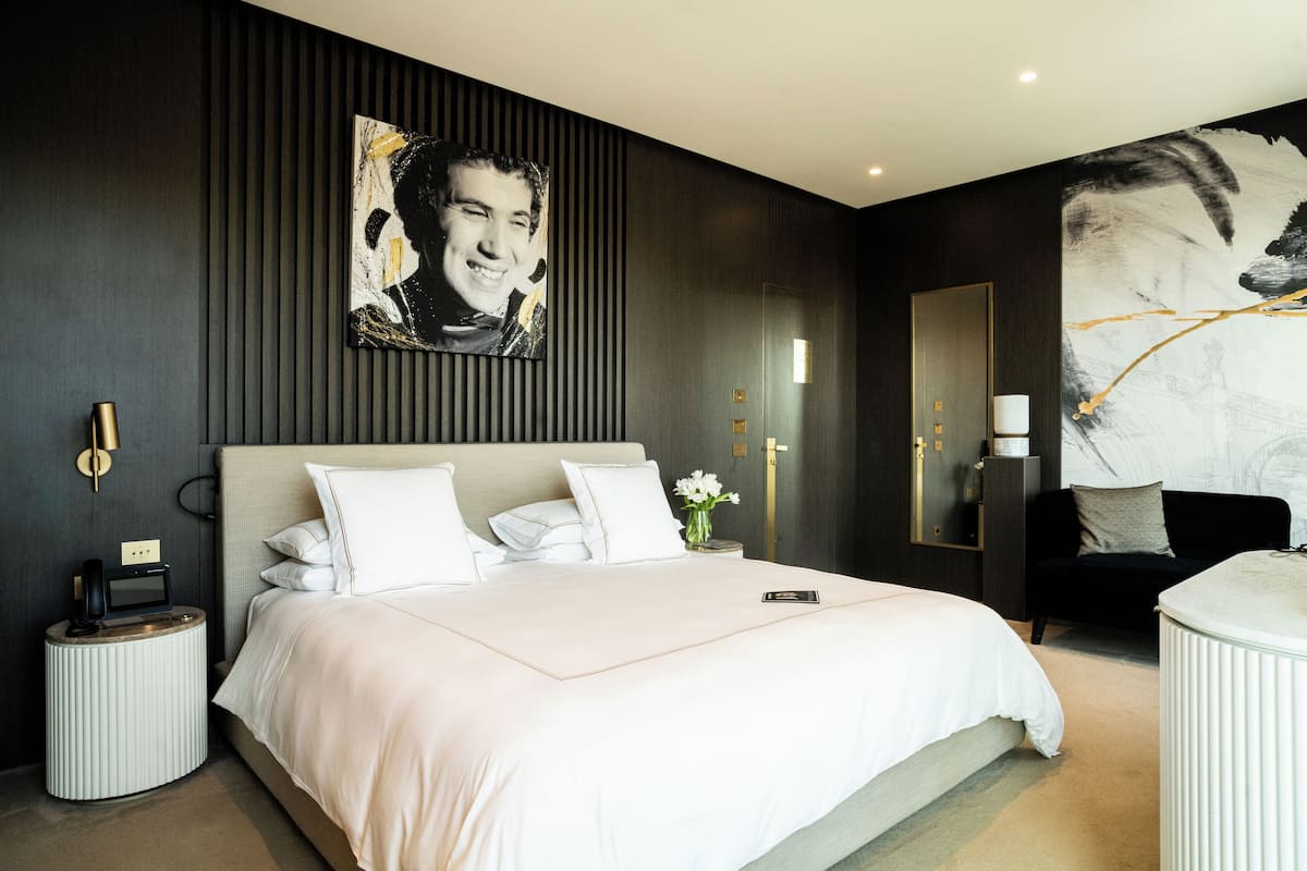 A modern hotel room with a large bed, a bedside table with a phone, a small side table, a mirror, and a wall with a large painting.
