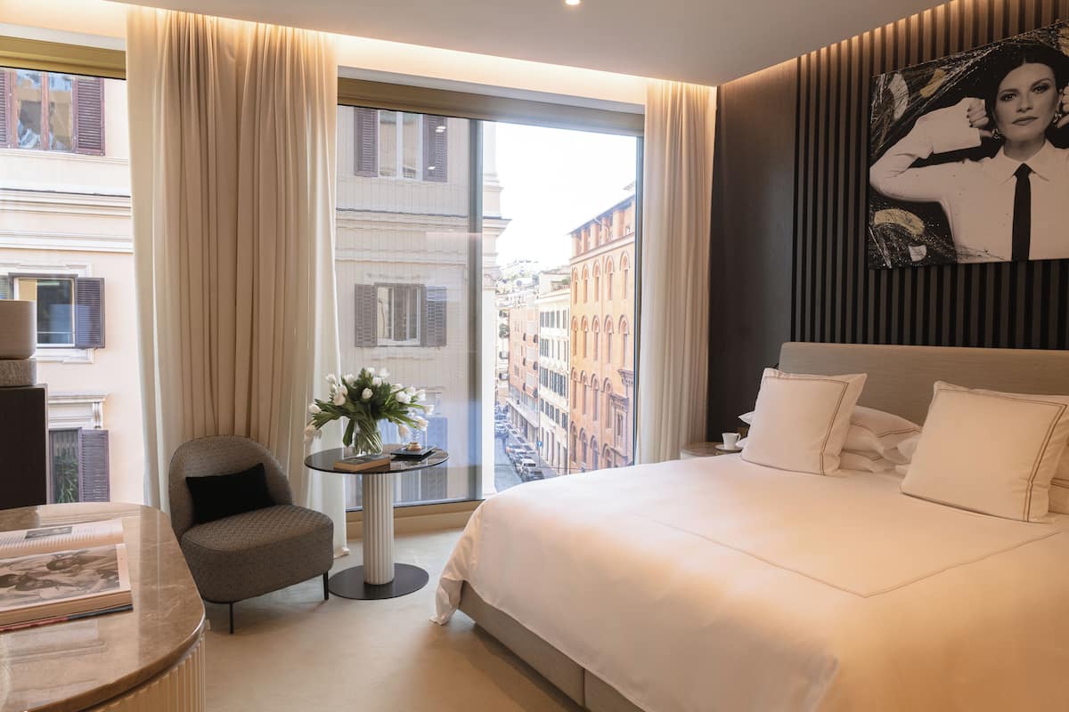 A hotel room with a large bed, a small round table with a vase, a chair, and a view of the city through the window.