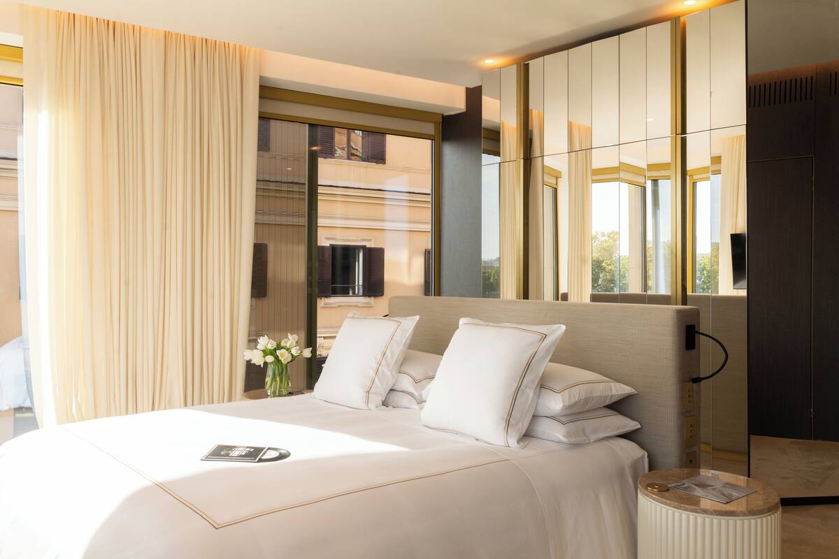 A modern hotel room with a large bed, a bedside table with a lamp, and a view of the city through large windows.