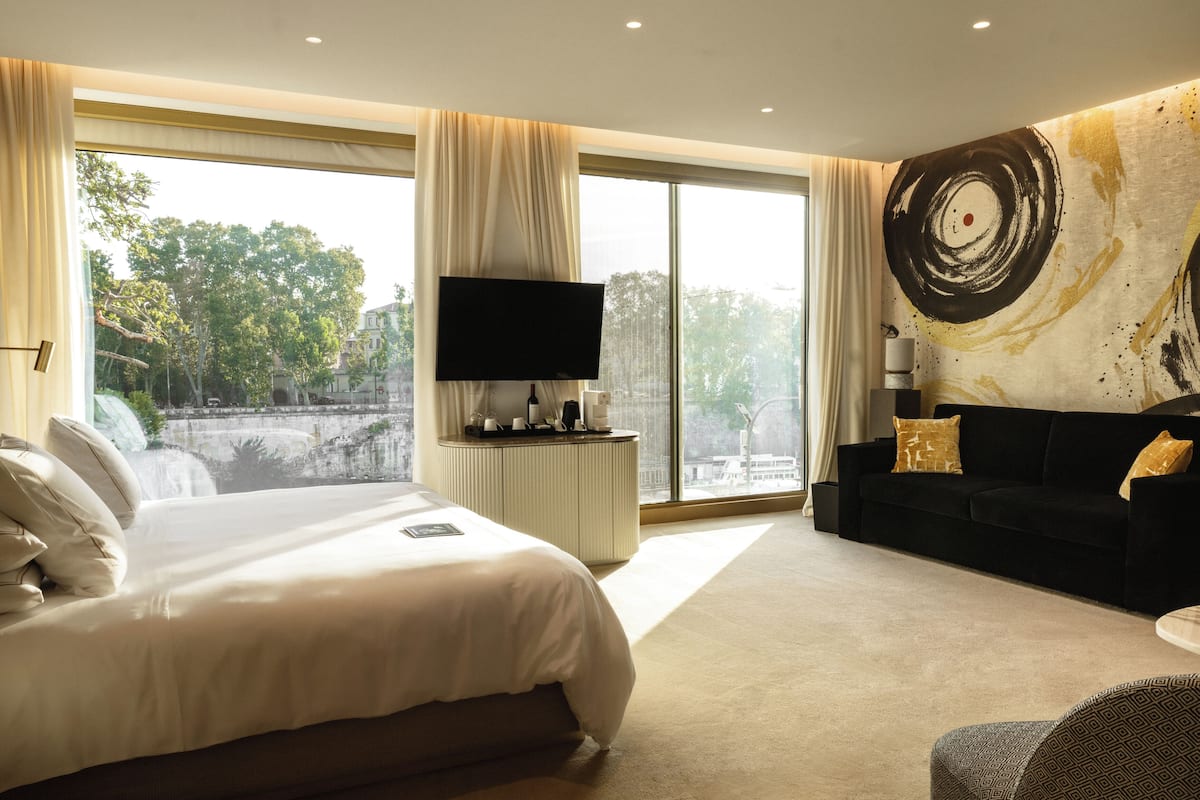 A hotel room with a large bed, a black sofa, a TV, and a view of trees and a bridge.