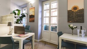 Un salón moderno con un sofá blanco, una mesa de comedor con sillas, una computadora portátil, una lámpara y un gran ventanal con persianas.