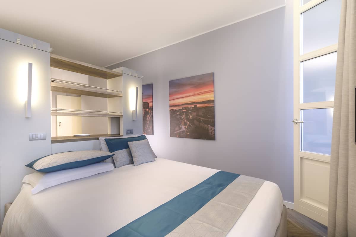 A modern hotel room with a large bed, built-in shelving, a cityscape painting, and a window with curtains.