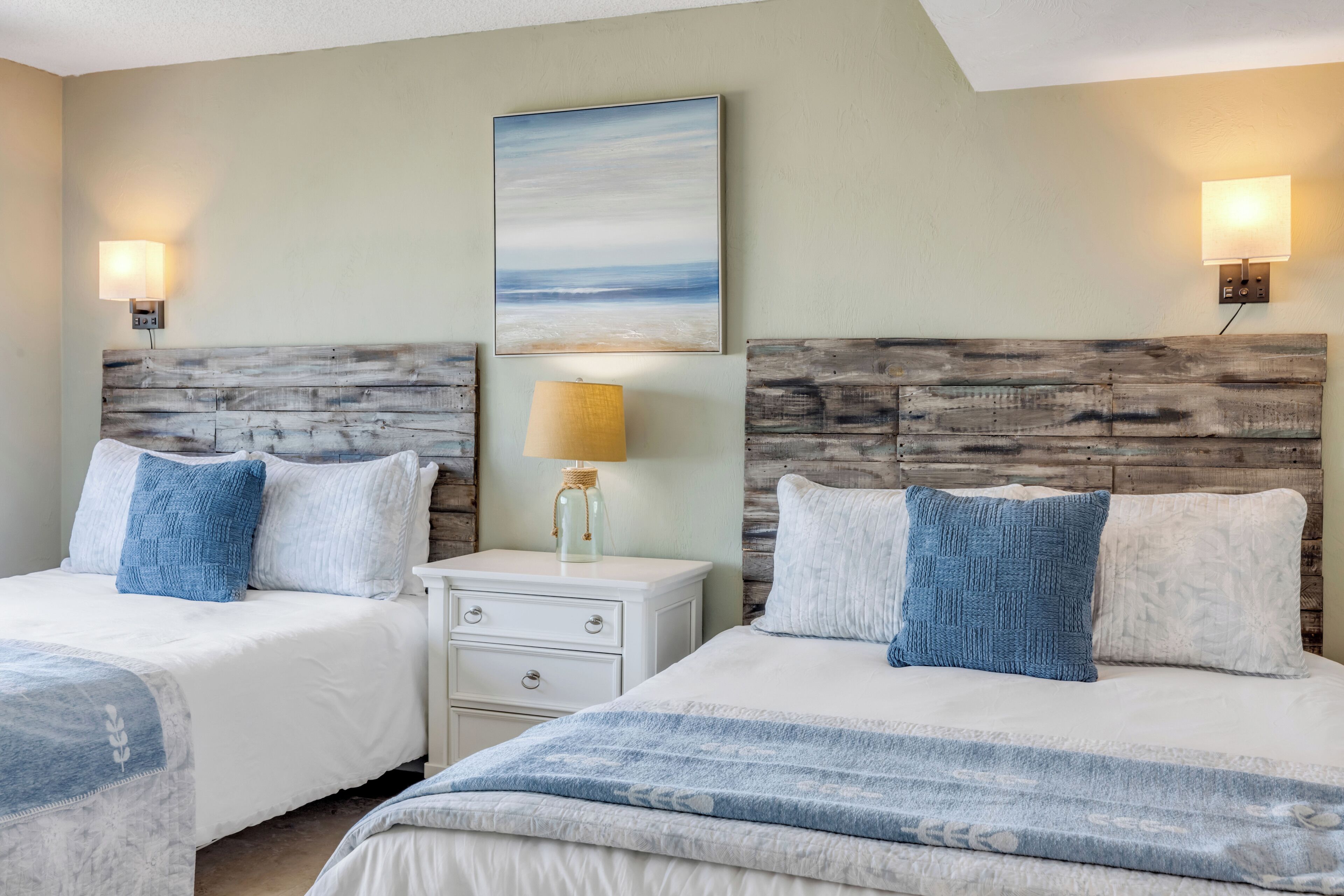 A hotel room with two beds, a nightstand, a lamp, a painting, and wall-mounted lights.