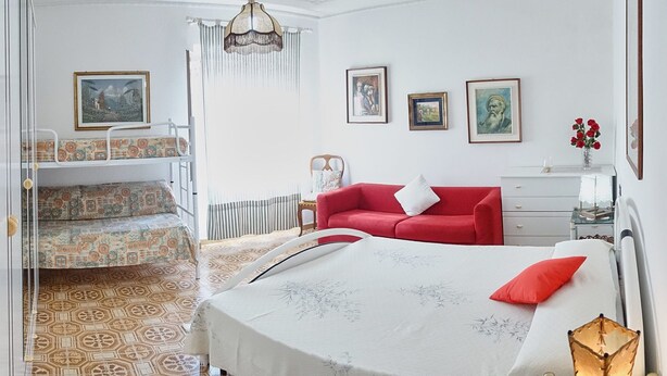 Ein Schlafzimmer mit einem Bett, einem roten Sofa, einem Sessel und einem kleinen Tisch. An der Wand hängen Bilder in Rahmen, und eine Chandelier ist angebracht.
