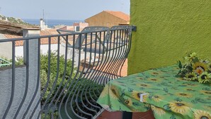 Ein Balkon mit einem Metallgeländer, einem Tisch mit einer Sonnenblumen-Tischdecke und Blick auf das Meer und Gebäude im Hintergrund.