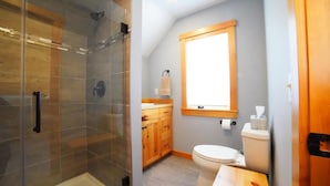 A bathroom with a glass shower enclosure, wooden cabinets, a toilet, and a window.