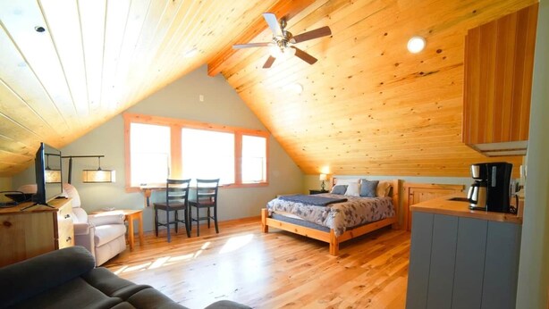 A cozy attic bedroom with a wooden ceiling, a bed, a desk, and a kitchenette.