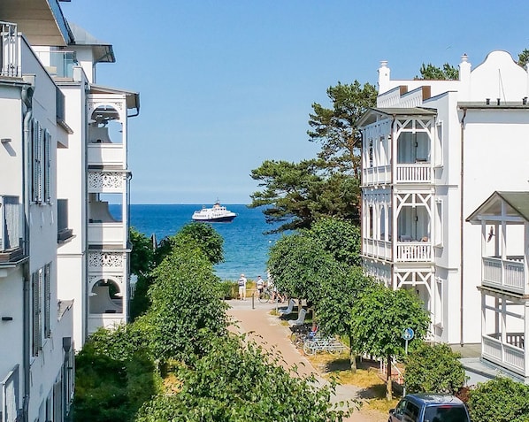 A coastal street with white buildings, a boat in the distance, and a pedestrian path.