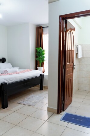 A bedroom with a bed, a potted plant, a wooden door, and a bathroom mat. - Honey Dew Stays (Athi River)