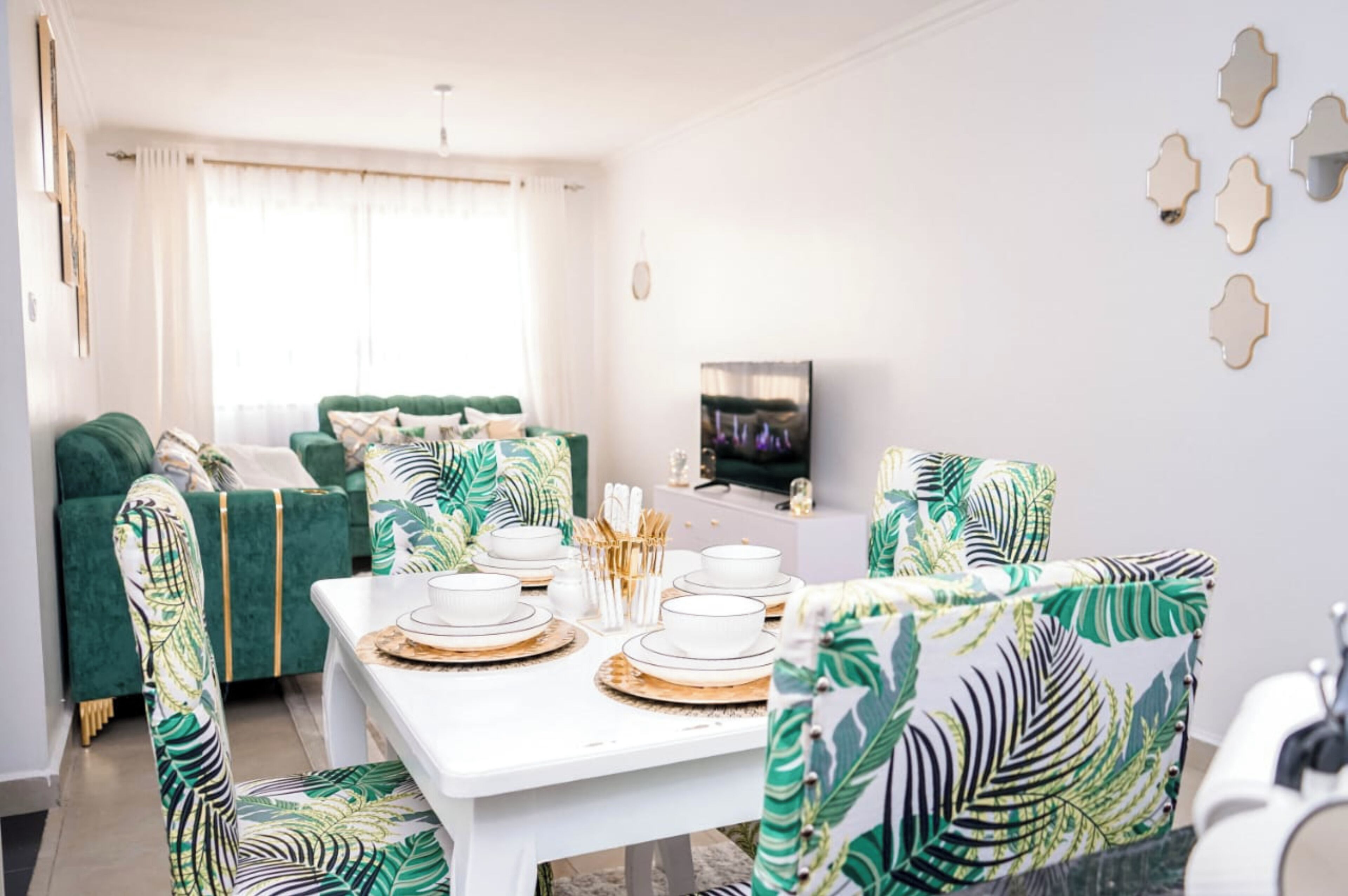 A dining area with a white table set for four, surrounded by green upholstered chairs with leaf patterns.