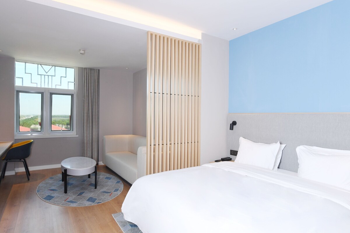A hotel room with a bed, a desk, a chair, and a view of the outdoors through a window.