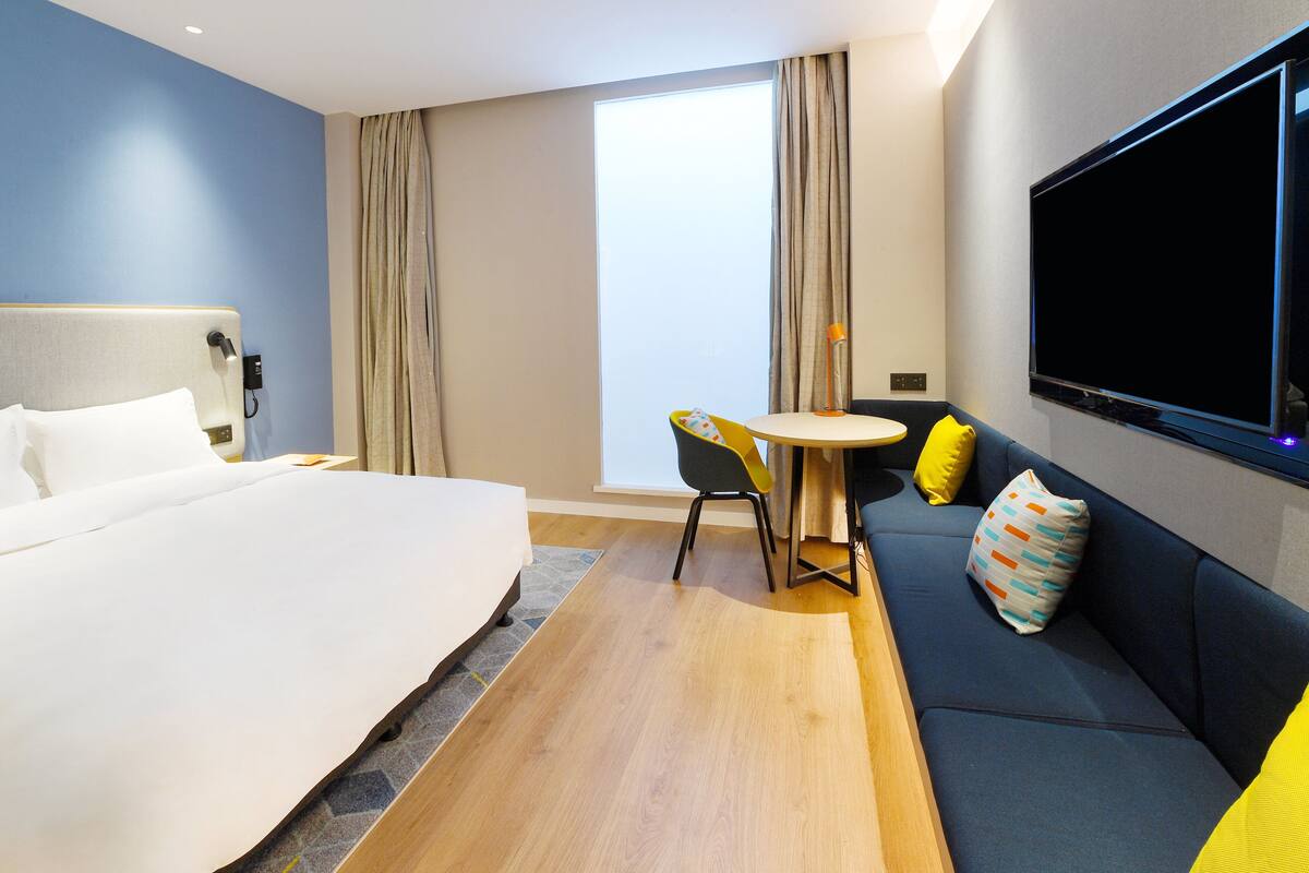 A hotel room with a bed, a sofa, a small table with a chair, a TV mounted on the wall, and a window with curtains.