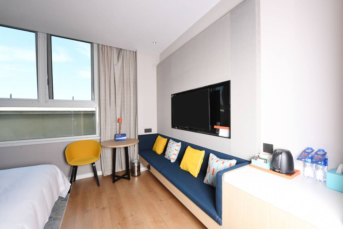 A hotel room with a bed, a sofa with yellow cushions, a small table, a TV, and a coffee machine.