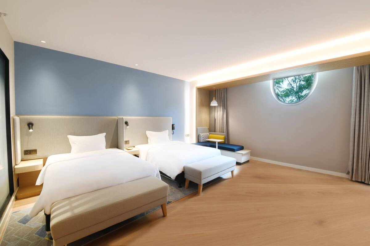 A modern hotel room with two beds, a small seating area, and a round window.