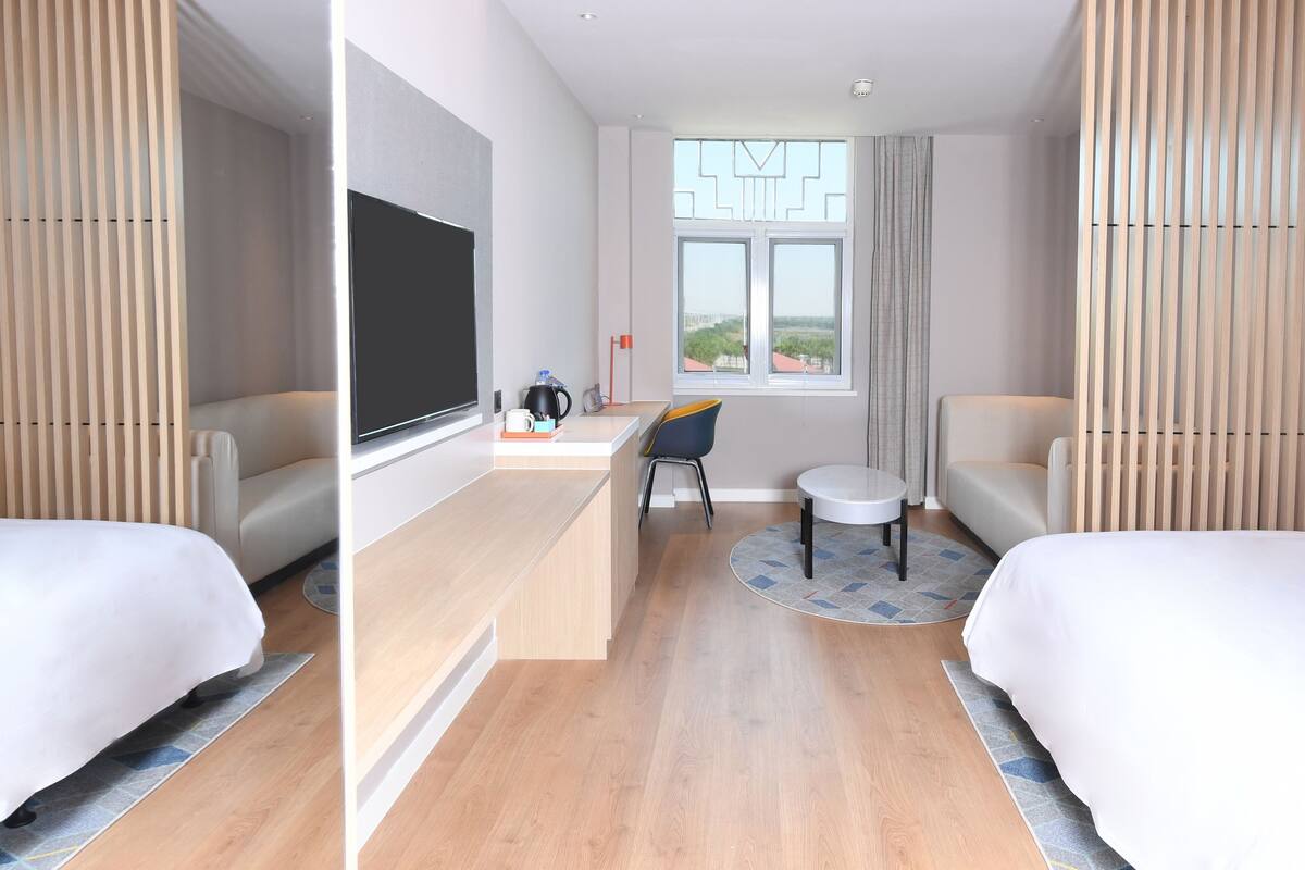 A hotel room with two beds, a desk, a chair, a TV, and a window with a city view.