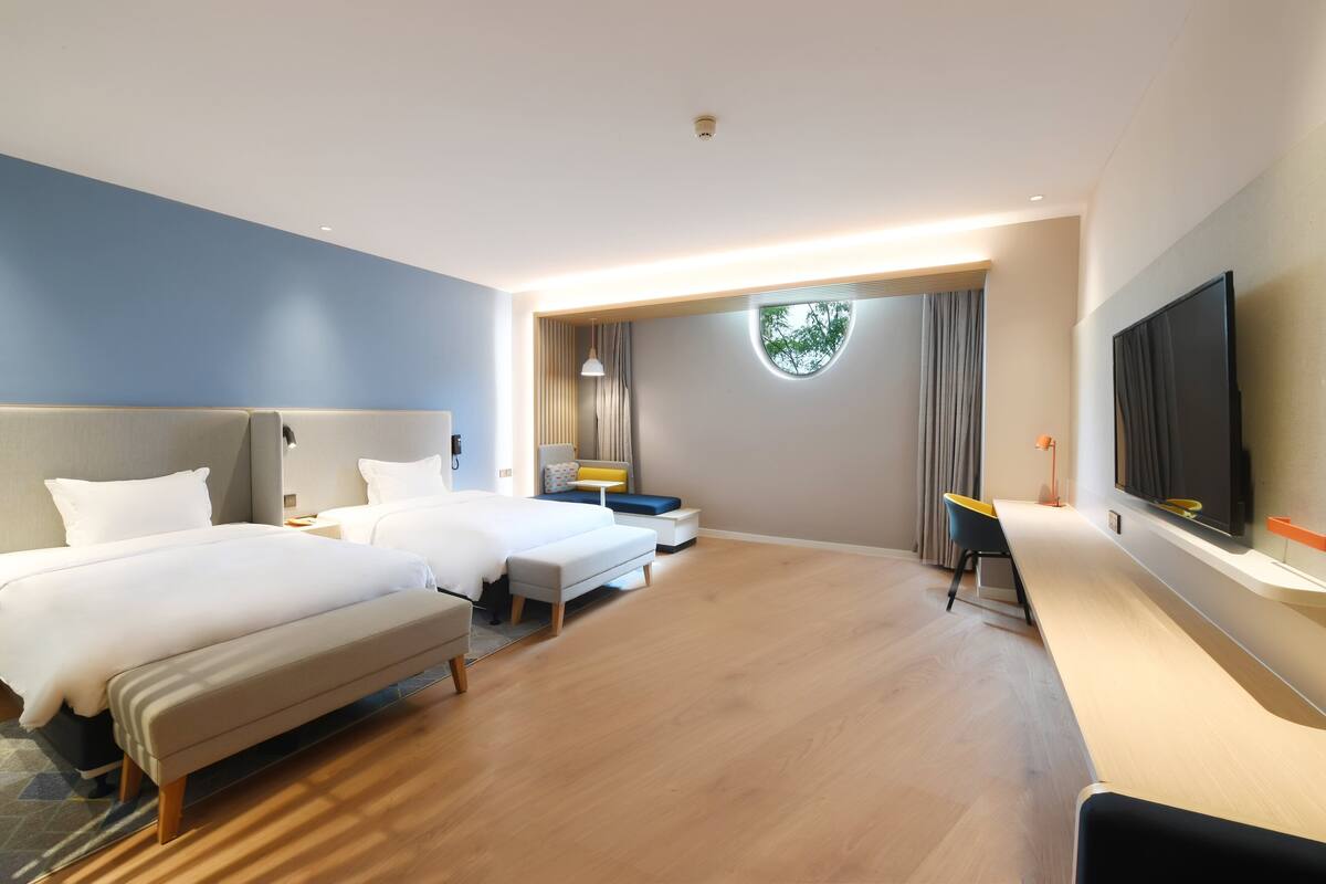 A hotel room with two beds, a desk, a TV, and a large window with curtains.