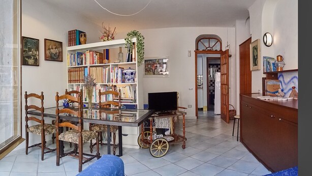 A living room with a glass dining table, wooden chairs, a bookshelf, a television, and a kitchen visible in the background. - Apartment (Santa Teresa Gallura)