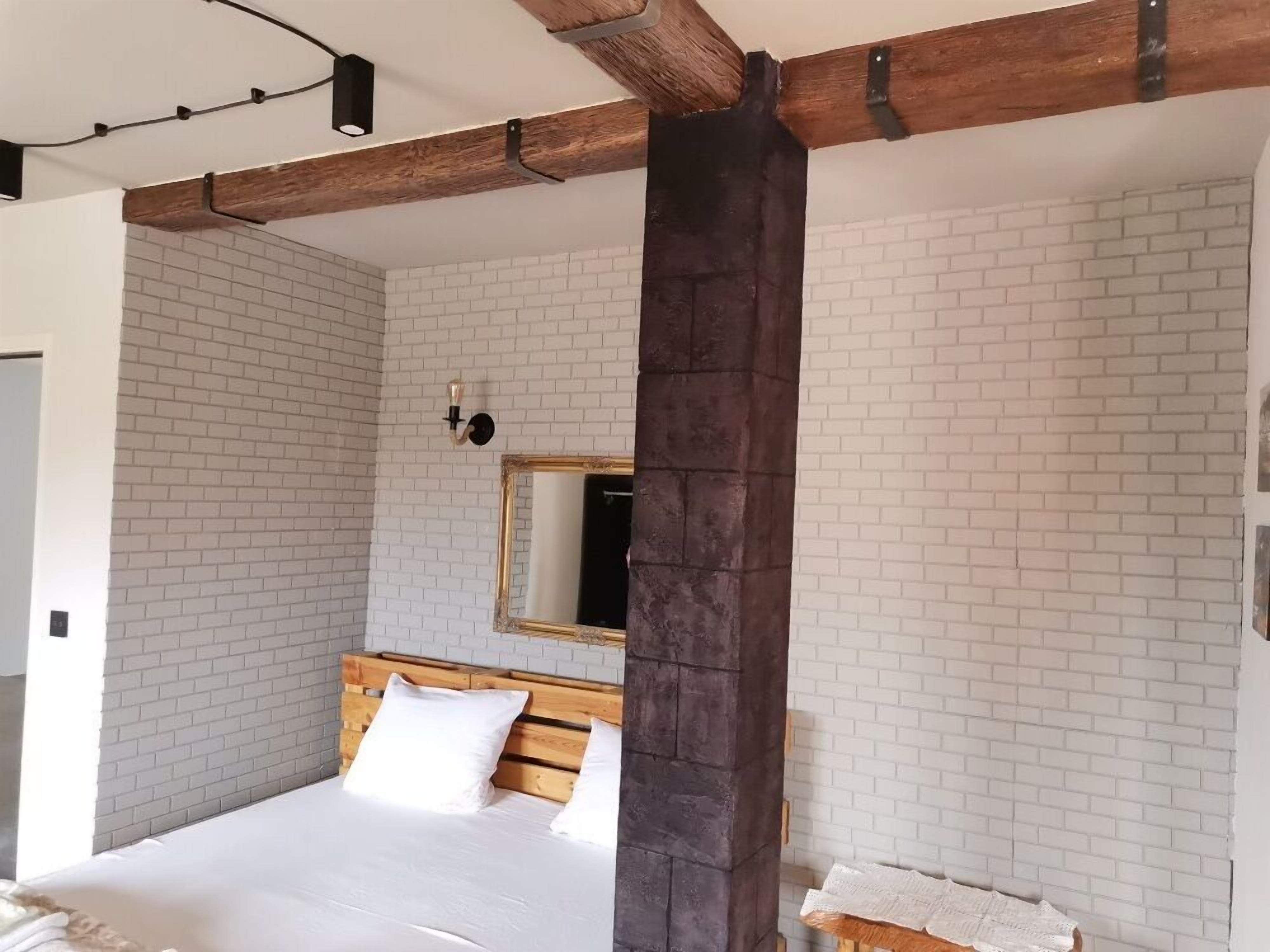 A bedroom with a wooden headboard, white brick walls, a large mirror, and a dark pillar.