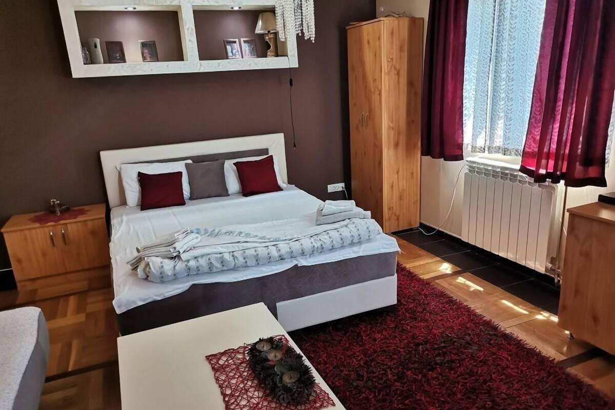 A bedroom with a bed, wooden nightstands, a wardrobe, a radiator, and a window with curtains.
