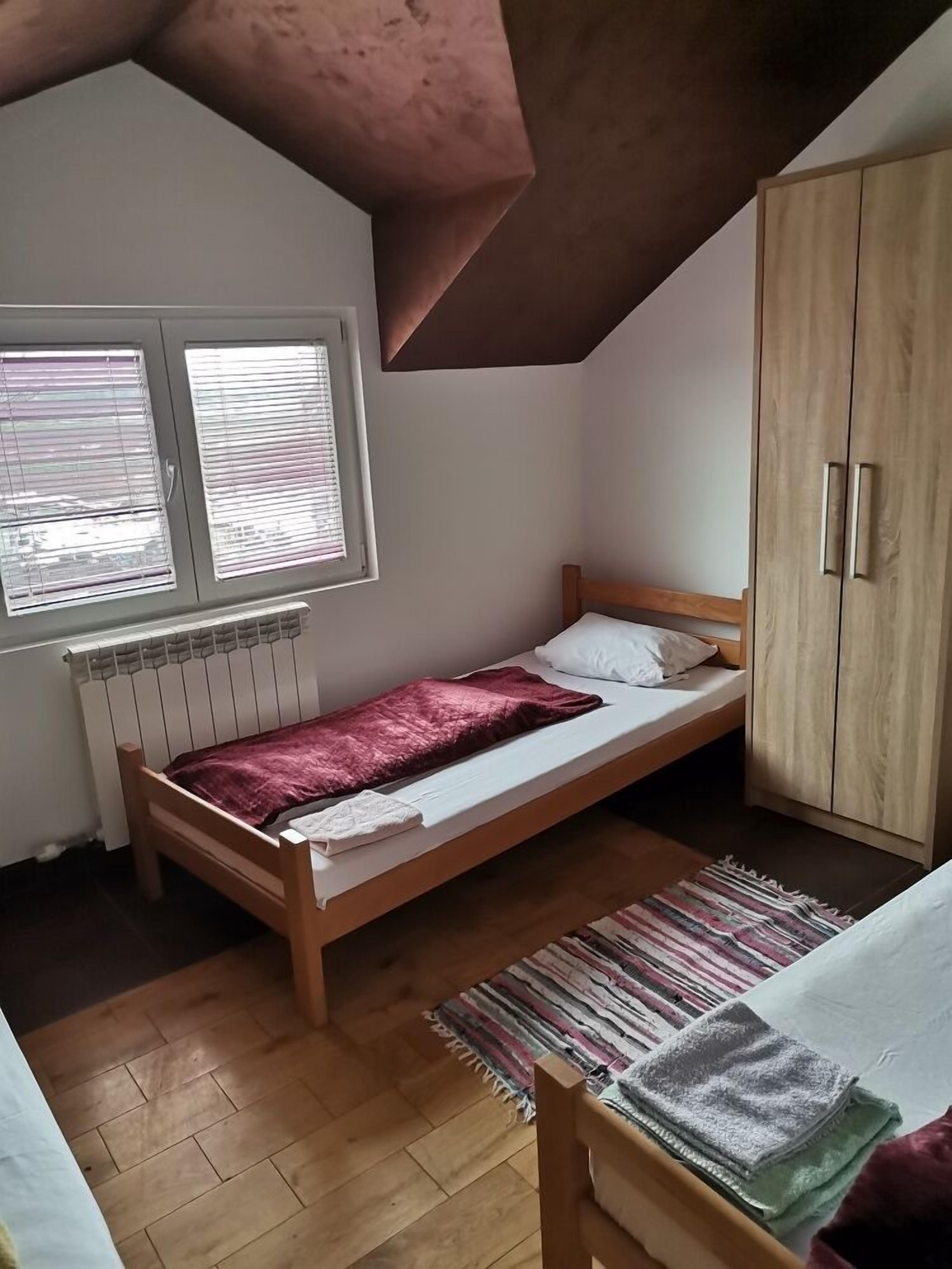 Ein kleines Zimmer mit einem Einzelbett, geneigter Decke, Holzboden, Heizkörper, Fenster mit Jalousien und einem Kleiderschrank.