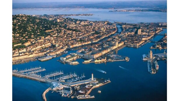 Vista aérea de uma cidade costeira com um porto de iates, área urbana densa e um farol imponente.