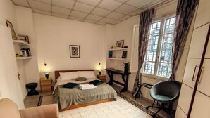 Ein Schlafzimmer mit Bett, Nachttischen, einem Sessel, einem Schreibtisch, einem Fenster mit Vorhängen und Regalen mit Büchern und Bildern.