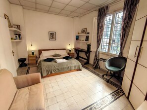 Een slaapkamer met een bed, nachtkastjes, een stoel, een bureau, een raam met gordijnen, en planken met boeken en foto’s.