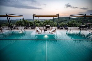 A rooftop infinity pool with lounge chairs and a table, surrounded by a wooden deck and a view of the landscape.