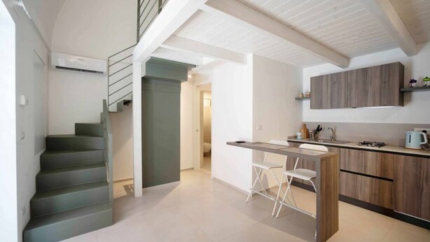 A modern kitchen with a built-in island, a staircase, and a view into another room. - WePuglia - Dimore Stefania - Suite Viuzza (Polignano a Mare)