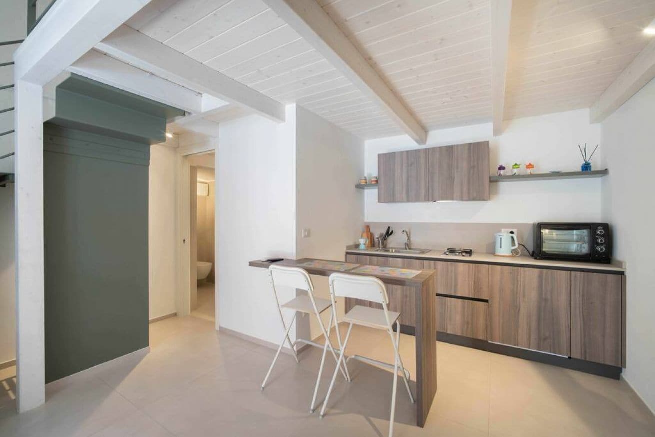 Una cocina moderna con encimera de madera, microondas integrado y un lavabo. Hay dos sillas blancas y una pequeña isla con una mesa.