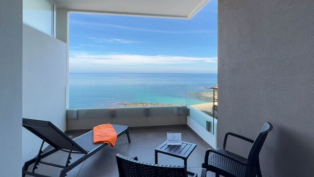 A balcony with a view of the sea, a lounge chair, a small table, and a sign.
