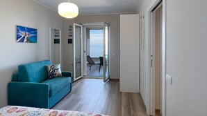 A modern room with a teal sofa, a bed with a floral bedspread, a pendant light, and a view to an outdoor seating area.