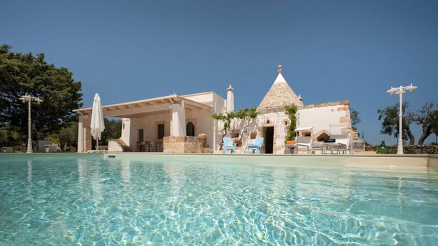 Área de piscina com água cristalina, um edifício com cúpula de pedra e espaço para sentar ao ar livre.