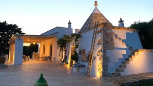 Um edifício de pedra com telhado cónico, uma escada e um pátio bem iluminado com mobiliário de exterior.