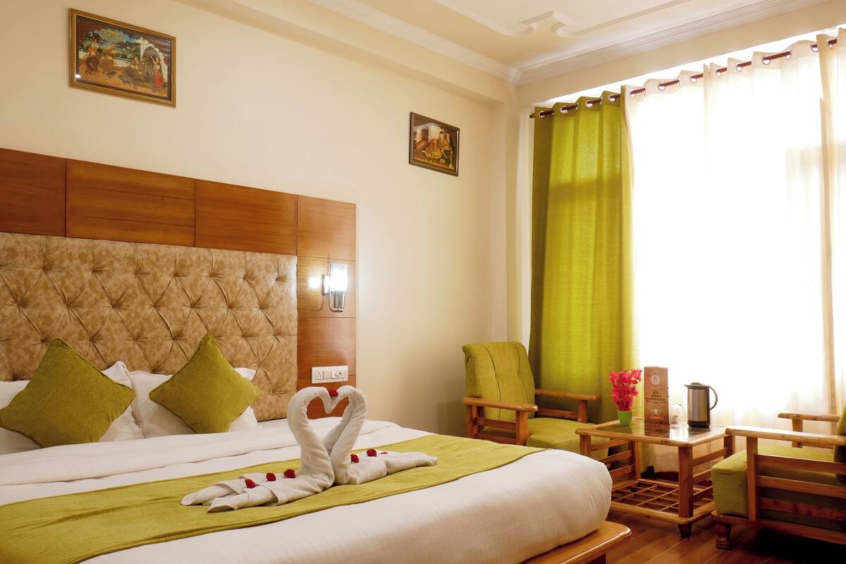 A hotel room with a large bed, a wooden headboard, a green chair, a small table with a pitcher, and a window with green curtains.