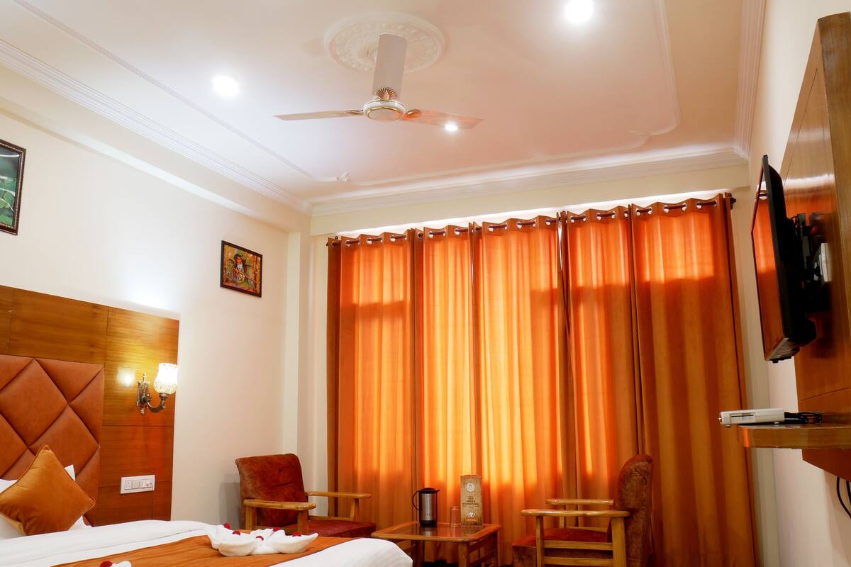 A hotel room with a bed, two wooden chairs, a small table with a lamp, a television mounted on the wall, and a large window with orange curtains.