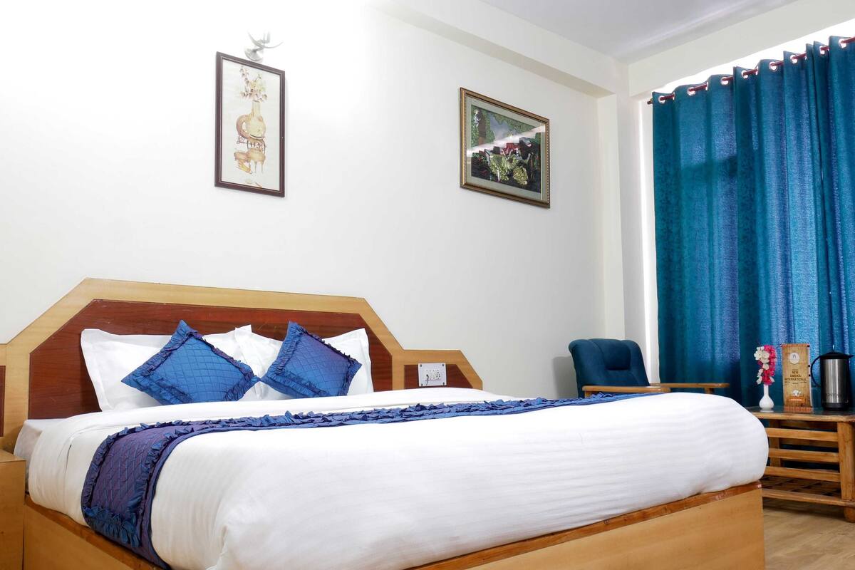 A neatly made bed with a wooden headboard, blue pillows, and a white comforter. There are two framed pictures on the wall, a blue chair, and a small table with a vase and a kettle.