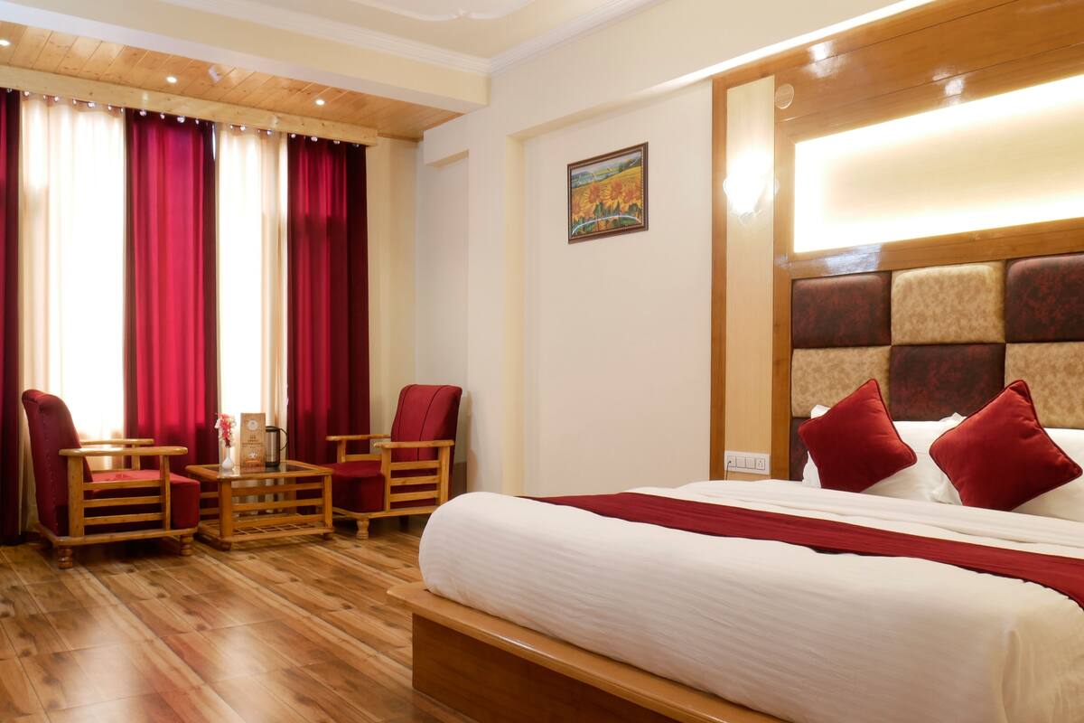 A hotel room with a wooden floor, a large bed with red pillows, a wooden headboard, a small table with a chair, and red curtains.