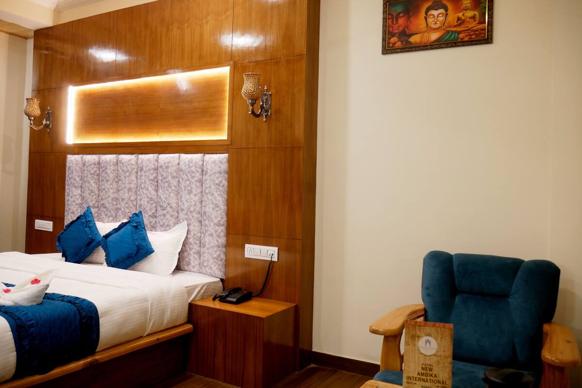 A hotel room with a wooden headboard, a bed with blue and white bedding, a bedside table with a lamp, a blue armchair, and a framed Buddha painting on the wall.
